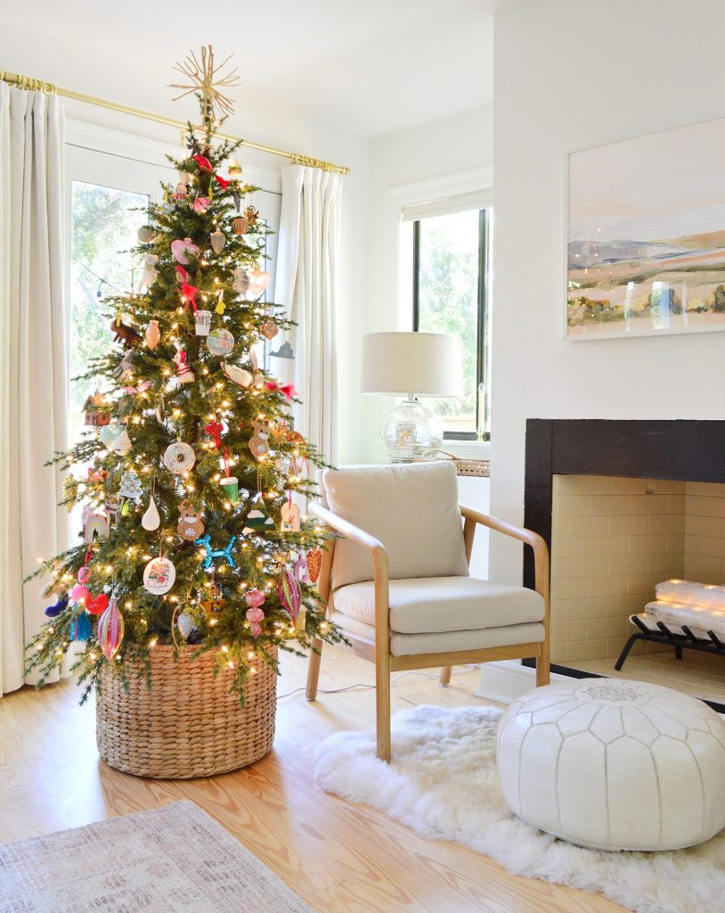 Slim Faux Christmas Tree In Woven Basket Next To Modern Chair And Fireplace