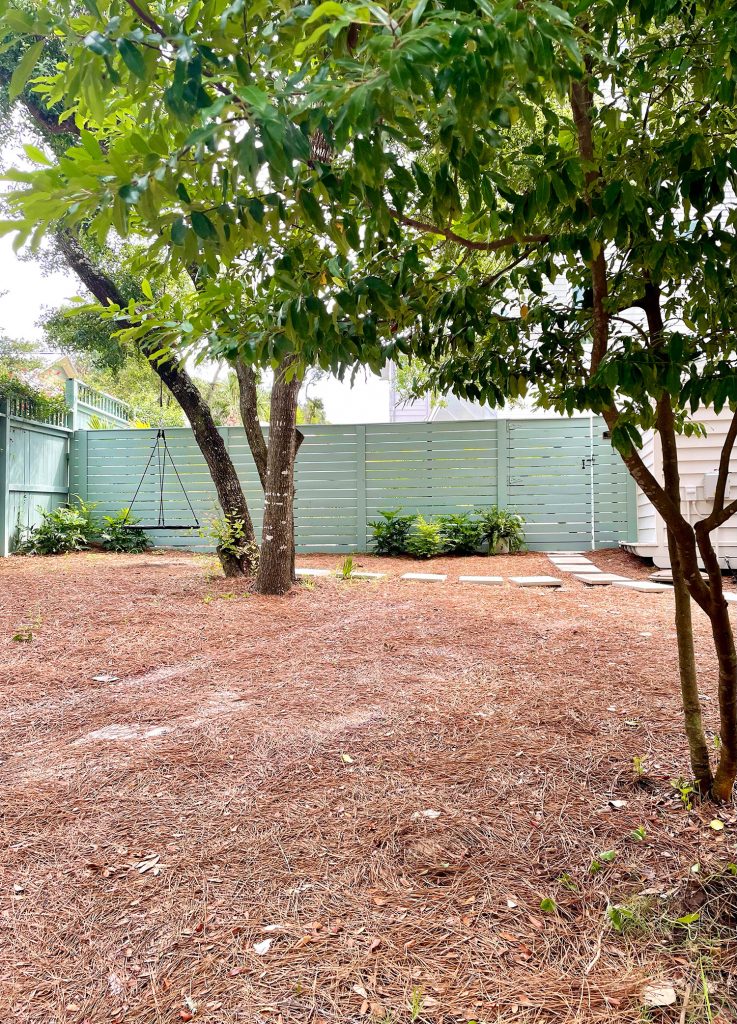 Large yard area with pine straw with green fence in background with tire swing on oak tree