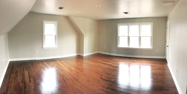 Wide angle panoramic photo of an empty bonus room with tan walls, wood floor, windows, and niche nook