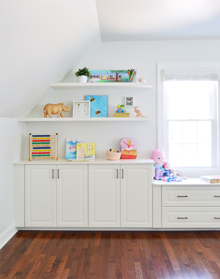 Two white floating shelves styled nicely with cheerful kids games and toys above white Ikea cabinets