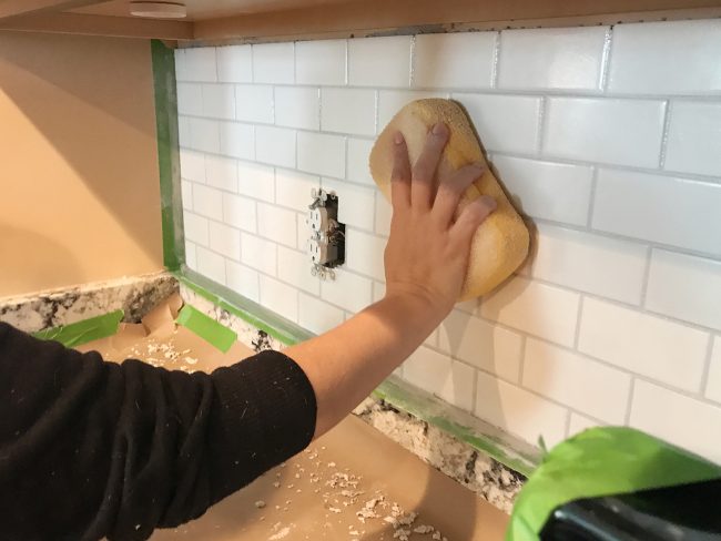 Sherry limpando o rejunte liso no backsplash da cozinha usando uma esponja úmida