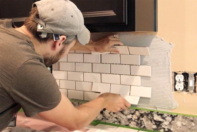 John placing white subway tile sheet onto mastic to set backsplash