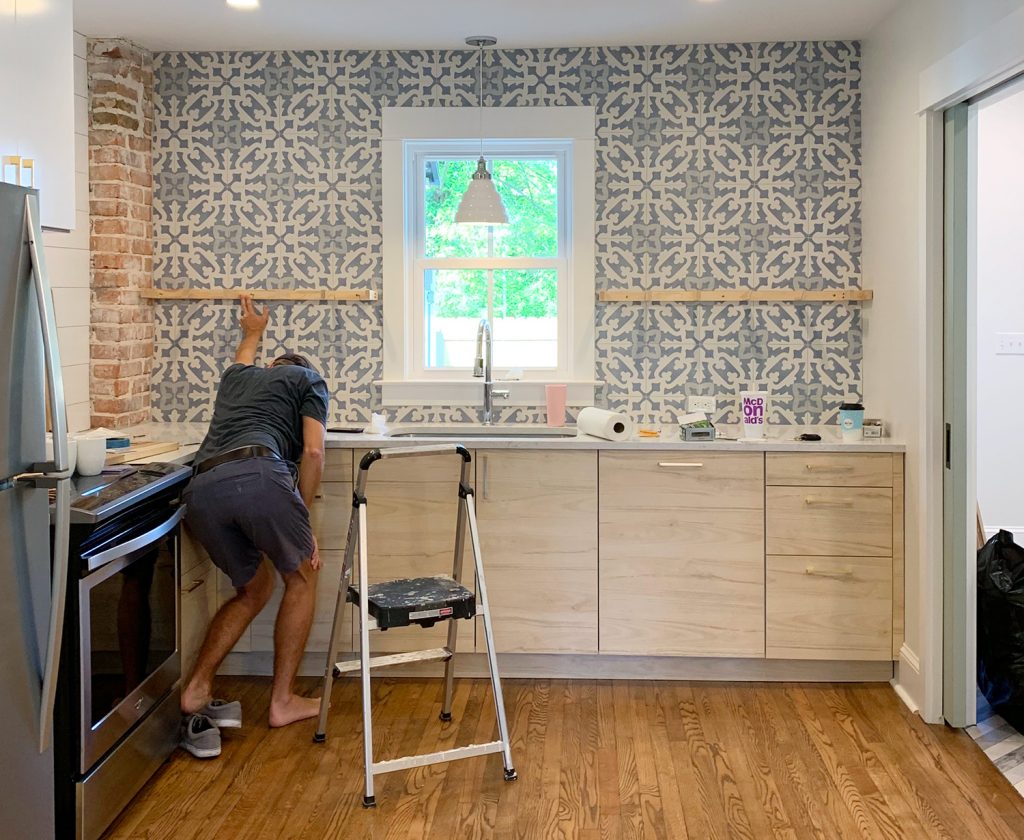 Man Crouching While Holding Kitchen Floating Shelf Bracket Against Tile For Placement