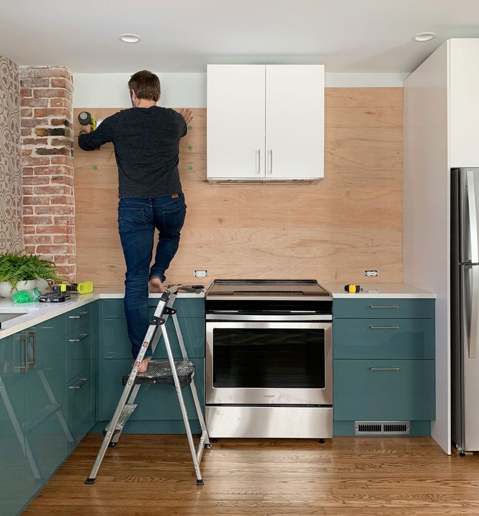 John em pé em uma escada pregando o backsplash com painéis de madeira em uma cozinha azul