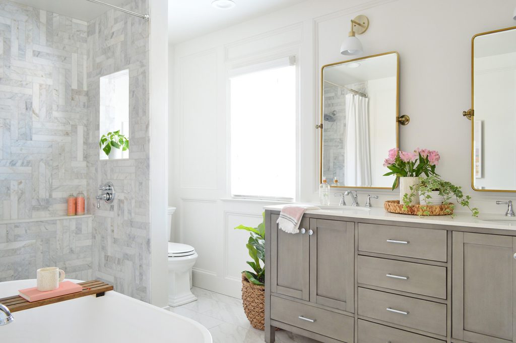 Marble Bathroom With Herringbone Tile Walk In Shower And Double Gray Vanity And Brass Pivot Mirrors