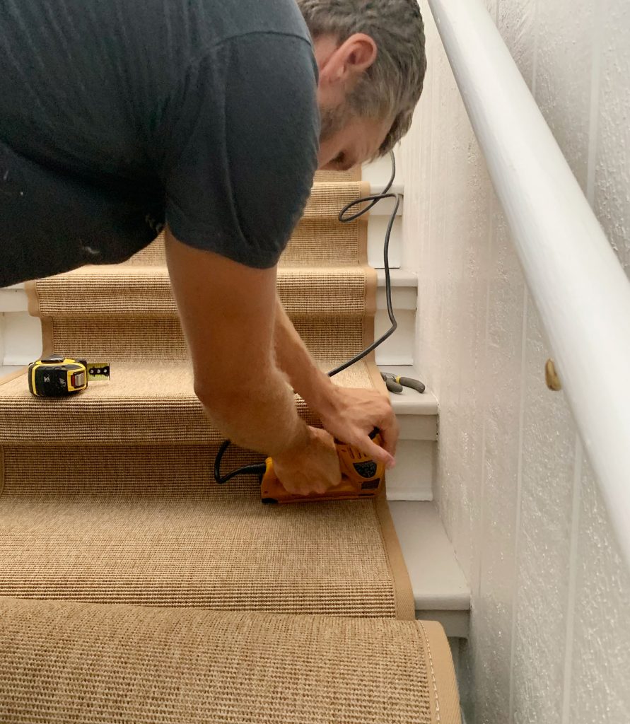 John usando uma pistola de grampos elétricos para colocar grampos no canto inferior da escada para fixar o corredor de sisal