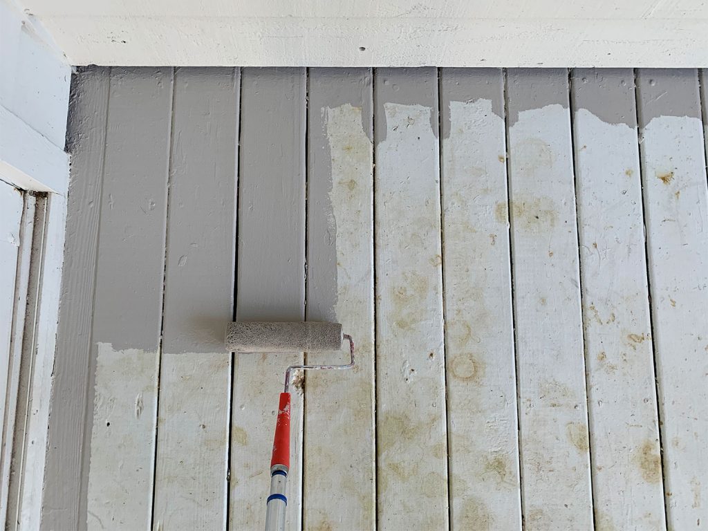 Roller applying gray porch paint to stained white porch floor