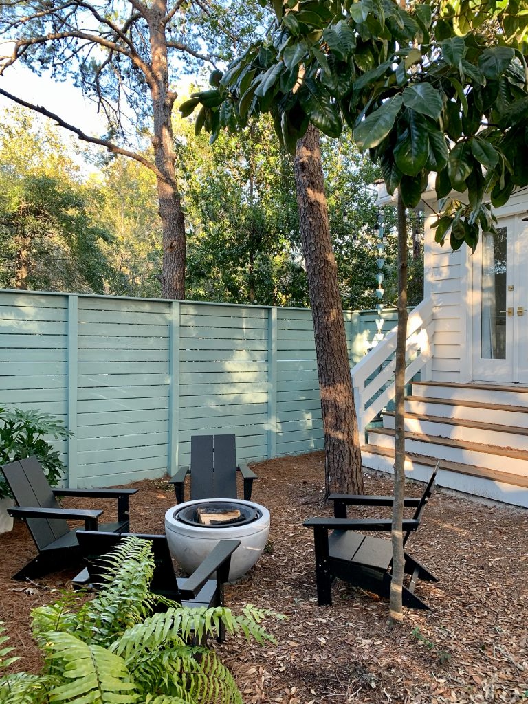 Trees around fenced side yard with black Adirondack chairs around fire pit