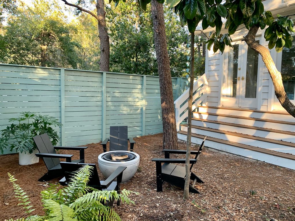 Firepit Area With Black Adirondack Chairs In Front Of Green Fence