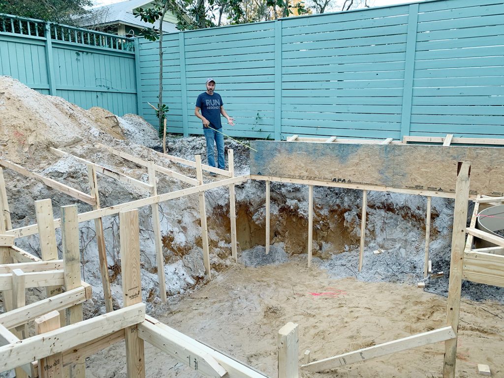 Buraco da piscina emoldurado com madeira e John ao fundo com fita métrica