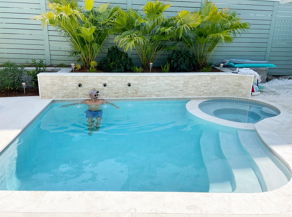 John em uma pequena piscina de forma livre contra um muro de contenção com detalhes em pedra e plantas de palmeiras