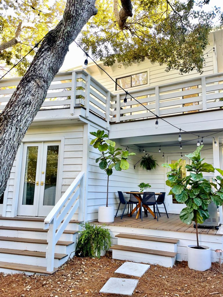 View of side porch with large oak tree in foreground with white house string lights and fig trees