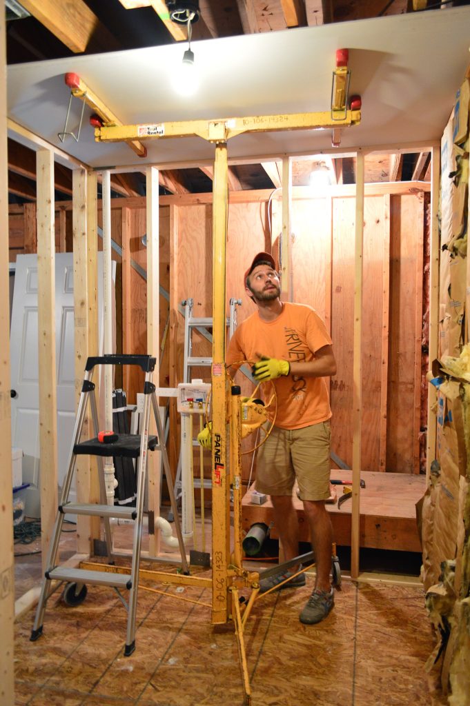 John using drywall lift to hold sheet of sheetrock to ceiling